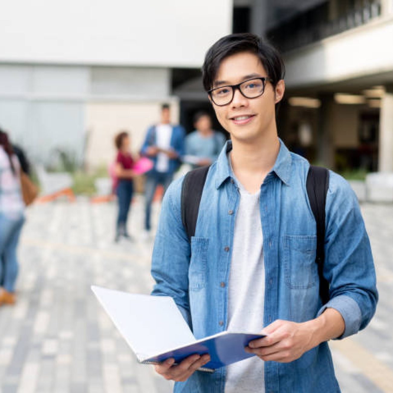 Portrait of happy exchange student facing camera smiling while holding his notebook at the university campus - Focus on foreground